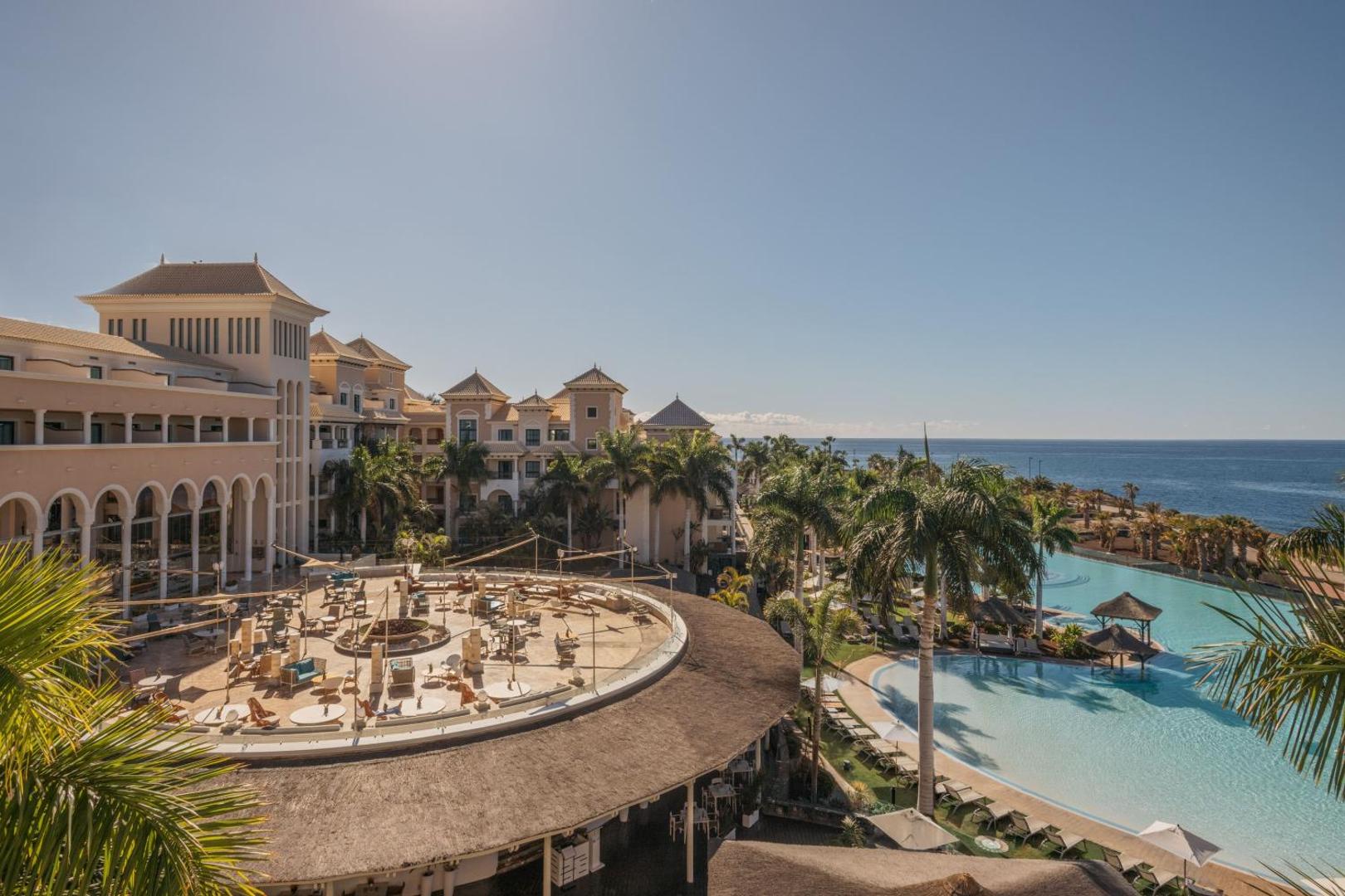 Gran Melia Palacio de Isora terase ar skatu uz okeānu un baseinu, palmas un atpūtas zona ar galdiņiem saulainā dienā