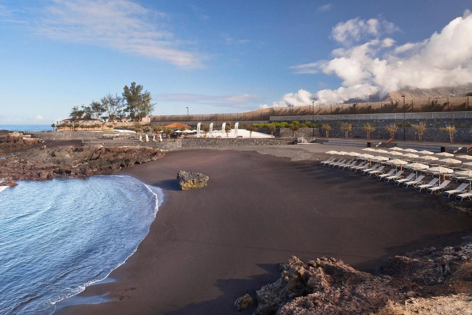 Gran Melia Palacio de Isora vulkānisko smilšu pludmale ar sauļošanās krēsliem, saulessargiem un klinšainu piekrasti Tenerifē.