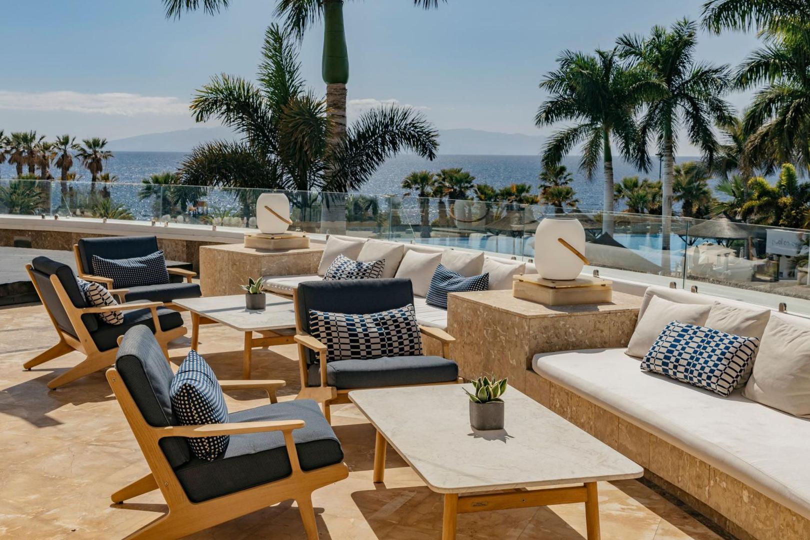 Viesnīcas Gran Melia Palacio de Isora terase ar modernām mēbelēm, dīvāniem un panorāmas skatu uz okeānu un palmām