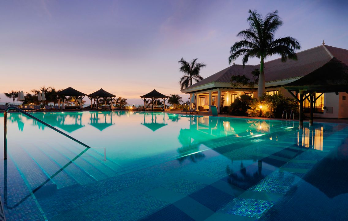 Gran Melia Palacio de Isora izgaismotais baseins un kūrorta ēka saulrieta laikā Tenerifē, radot romantisku atmosfēru.