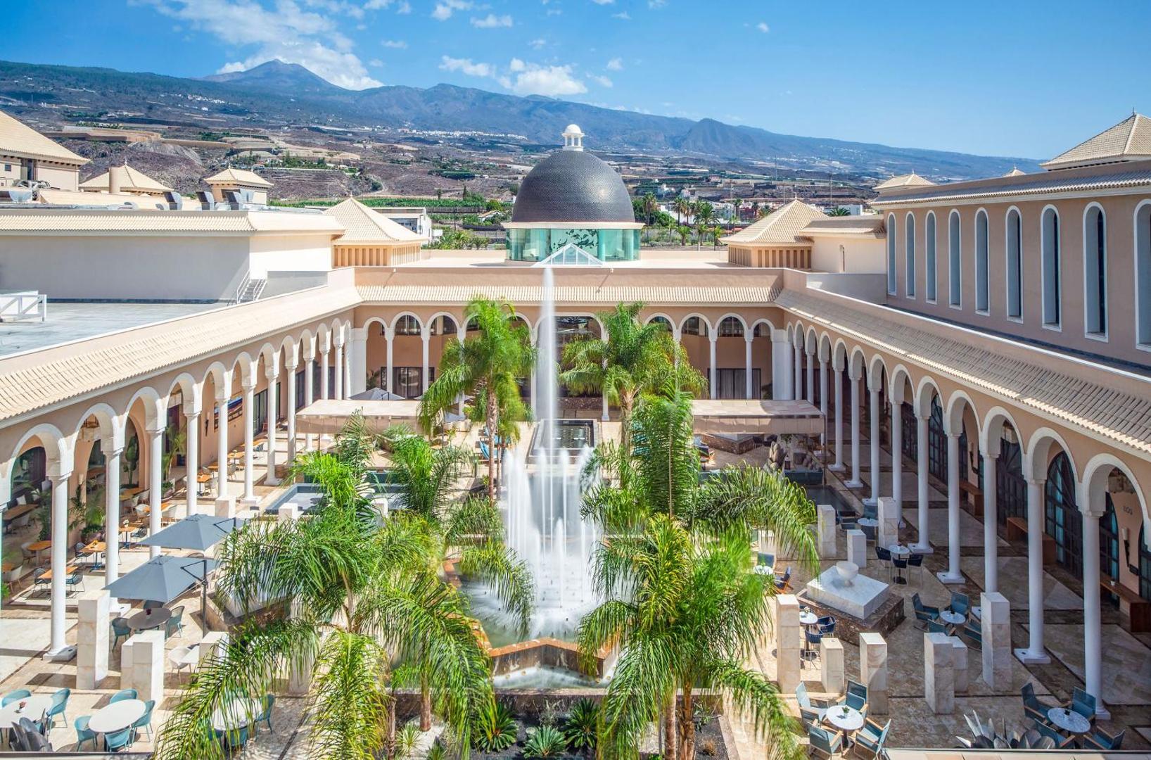 Gran Melia Palacio de Isora centrālais pagalms ar augstu strūklaku, palmām un skatu uz Teides kalnu fonā skaidrās debesīs