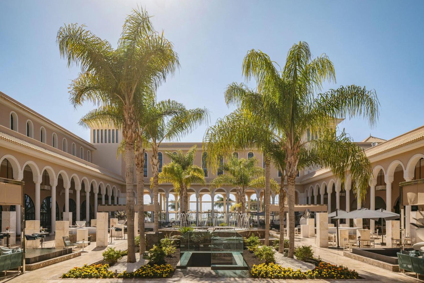 Gran Melia Palacio de Isora iekšpagalms ar palmām, strūklakām un klasisku arhitektūru, atpūtas zona ar krēsliem un saulessargiem