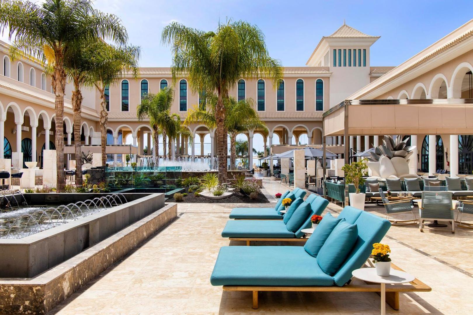 Gran Melia Palacio de Isora āra terase ar strūklakām, palmām un tirkīza krāsas atpūtas krēsliem pie kūrorta galvenās ēkas