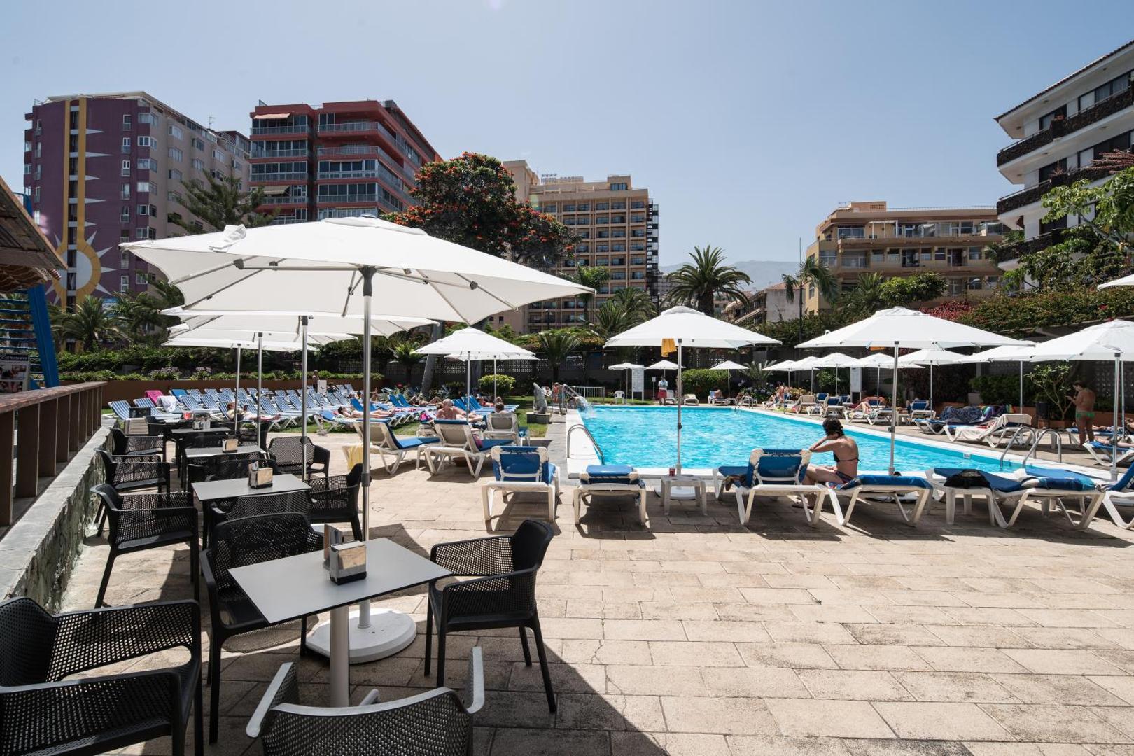 Catalonia Las Vegas terase ar galdiņiem un krēsliem pie baseina zonas, apkārt pilsētas apbūve un palmas Tenerifē