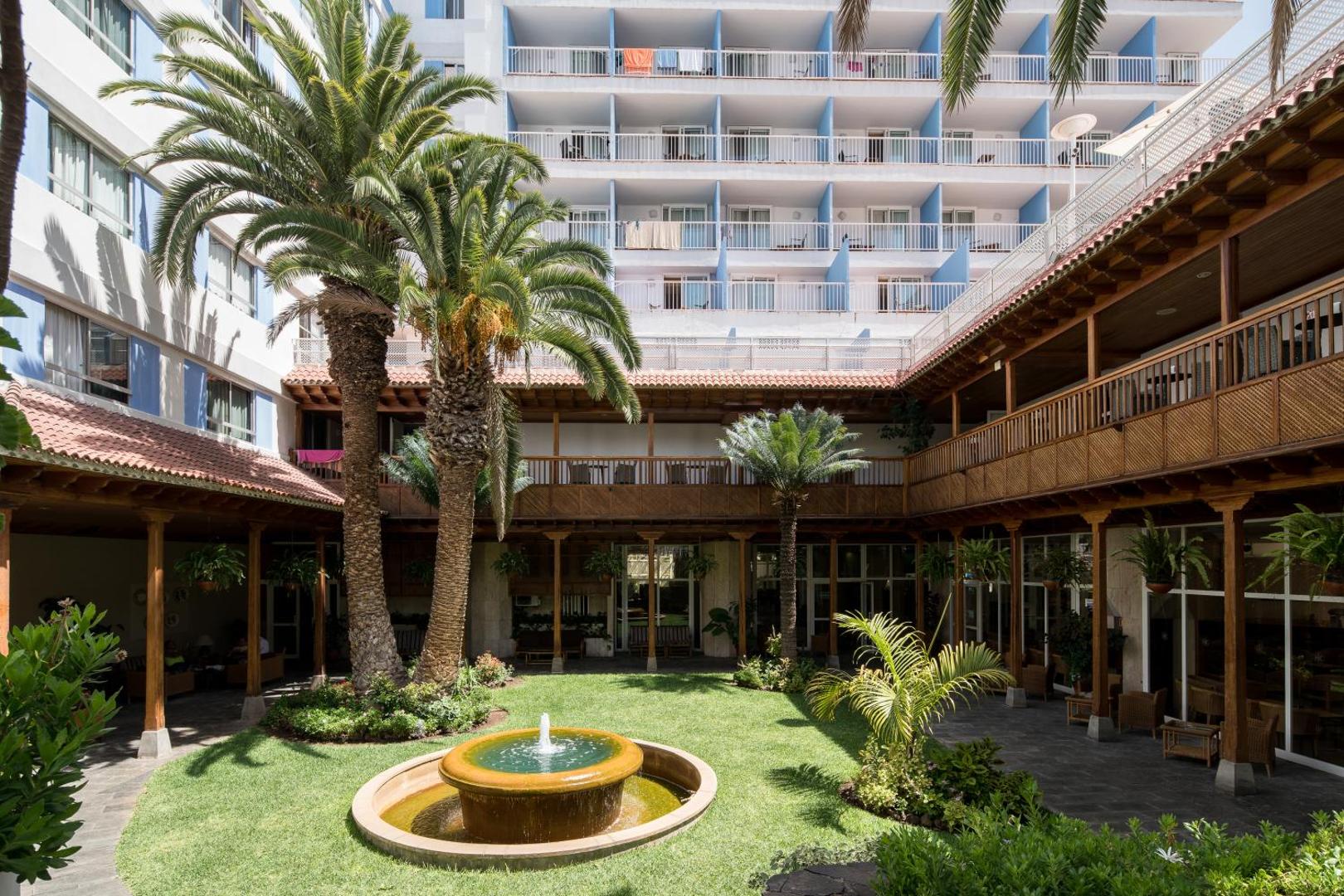 Catalonia Las Vegas iekšpagalms ar tradicionālu koka arhitektūru, zaļu zālienu, nelielu strūklaku un palmām Tenerifes kūrortā