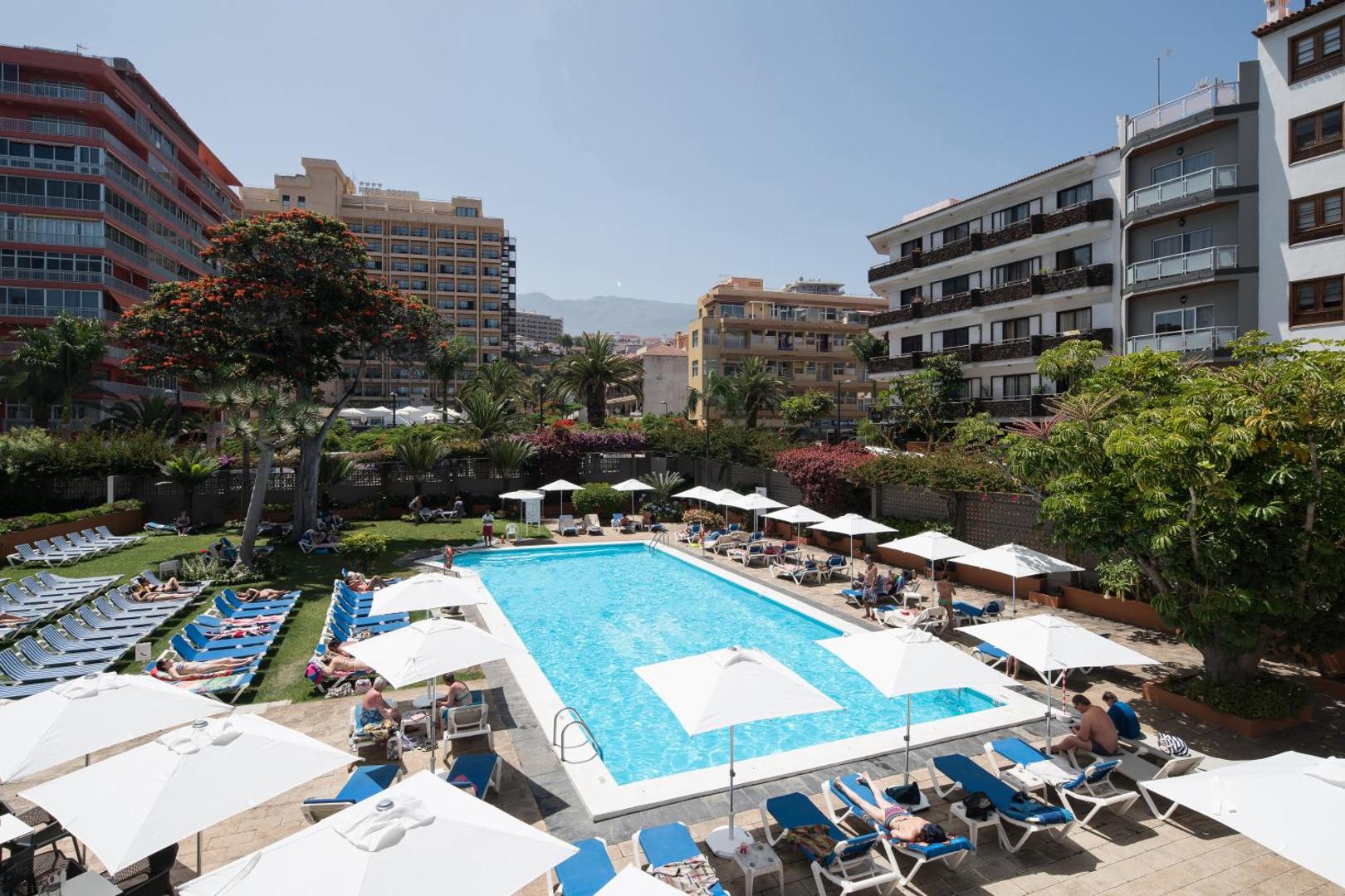 Viesnīcas Catalonia Las Vegas baseina zona ar zālāju, sauļošanās krēsliem un palmām saulainā dienā Tenerifes piekrastē