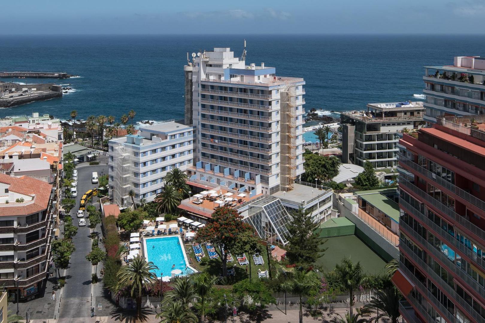 Panorāmas skats uz viesnīcu Catalonia Las Vegas pie okeāna krasta Puerto de la Krusā, Tenerifē — redzams baseins un pilsētas ainava
