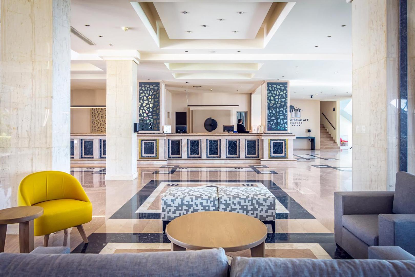 Kresten Palace Hotel vestibils ar modernām mēbelēm, dzeltenu atpūtas krēslu un skatu uz galveno reģistratūras zonu Rodas kūrortā