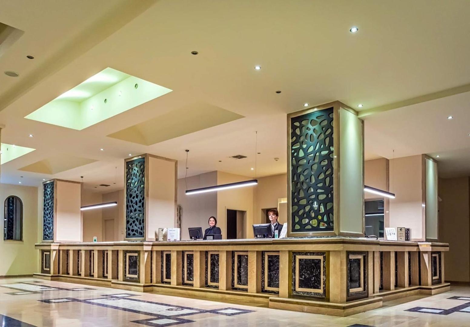 Kresten Palace Hotel reģistratūra Rodas salā ar plašu marmora leti, modernu apgaismojumu un viesmīlīgu personālu gaišā viesnīcas vestibilā