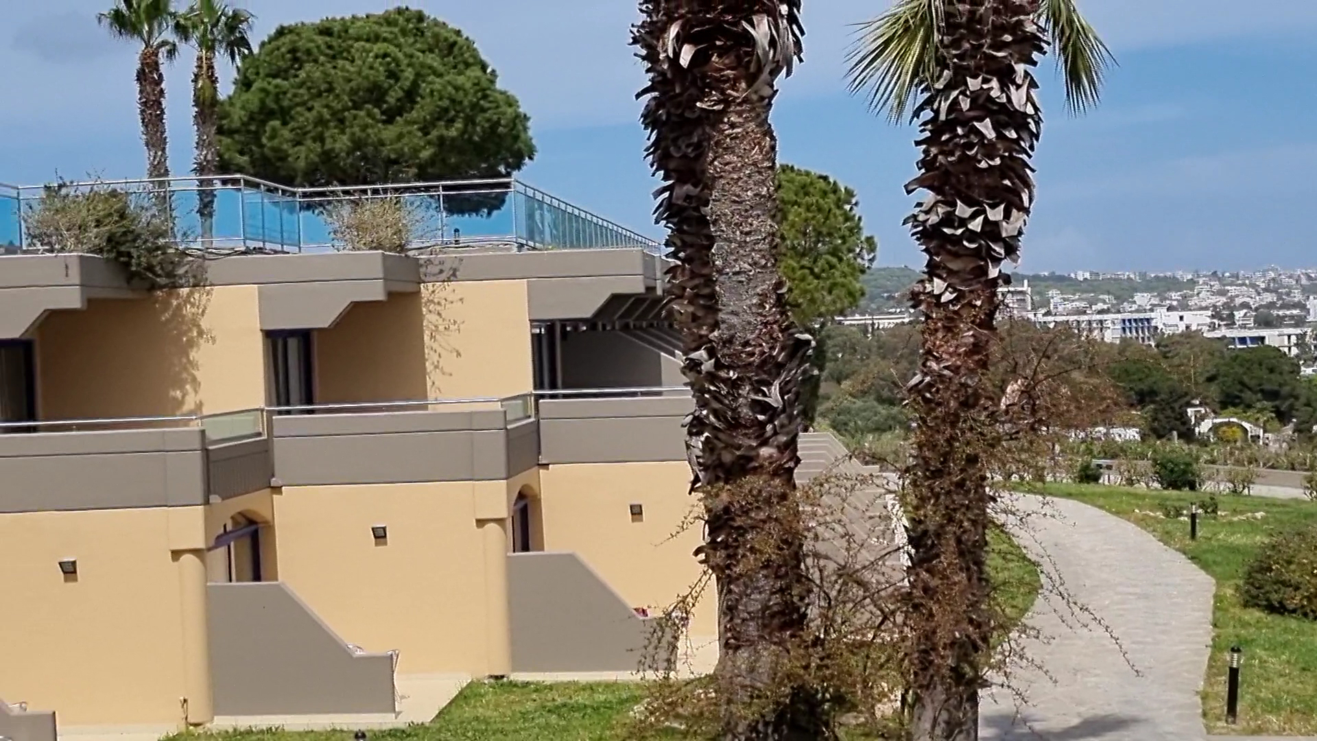 Kresten Palace Hotel dārza skats ar palmām un gājēju celiņu — viesnīcas numuru balkoni un skats uz pilsētu tālumā