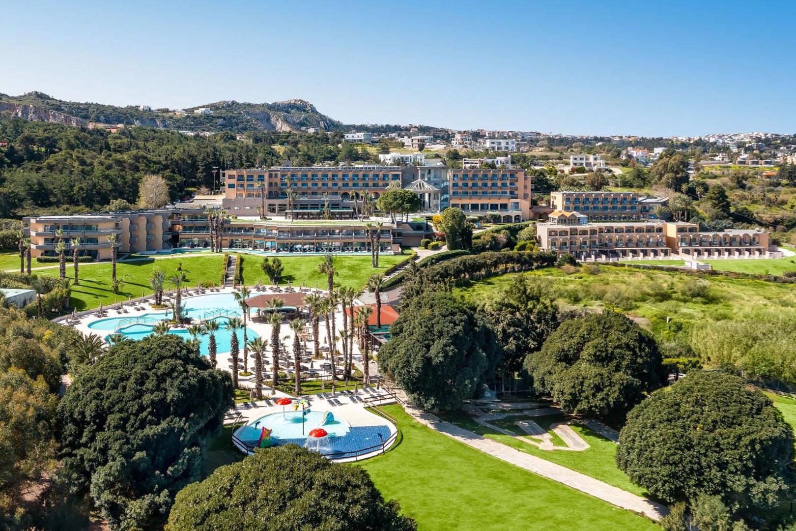Kresten Palace Hotel teritorijas skats ar bērnu baseinu un galveno ēku Rodas salā — kalnains fons un plašas atpūtas zonas