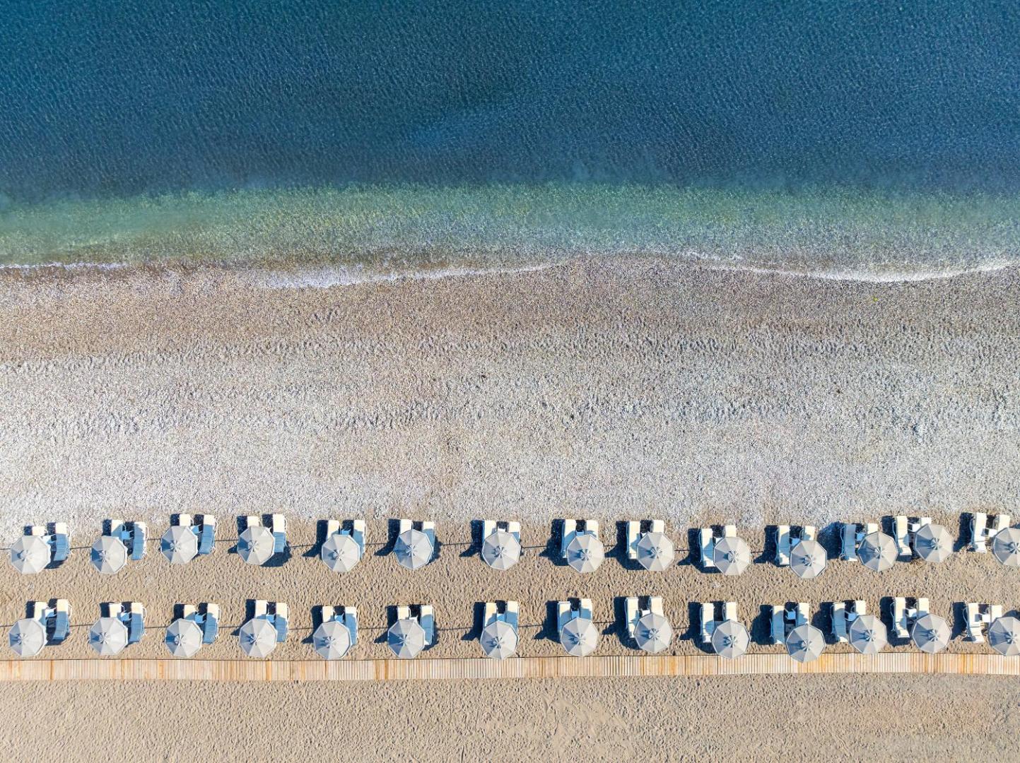 Sun Beach Resort pludmales josla no putna lidojuma — simetriskas saulessargu rindas uz smilšu un oļu seguma pie dzidra ūdens