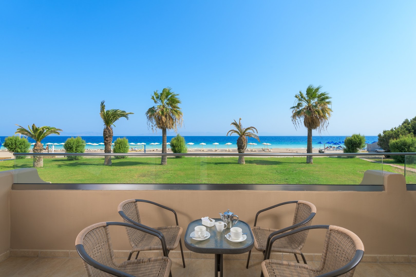 Sun Beach Resort numura balkons ar skatu uz jūru un palmām Rodas salā, Grieķijā — kafijas galdiņš ar krēsliem un pludmales panorāma