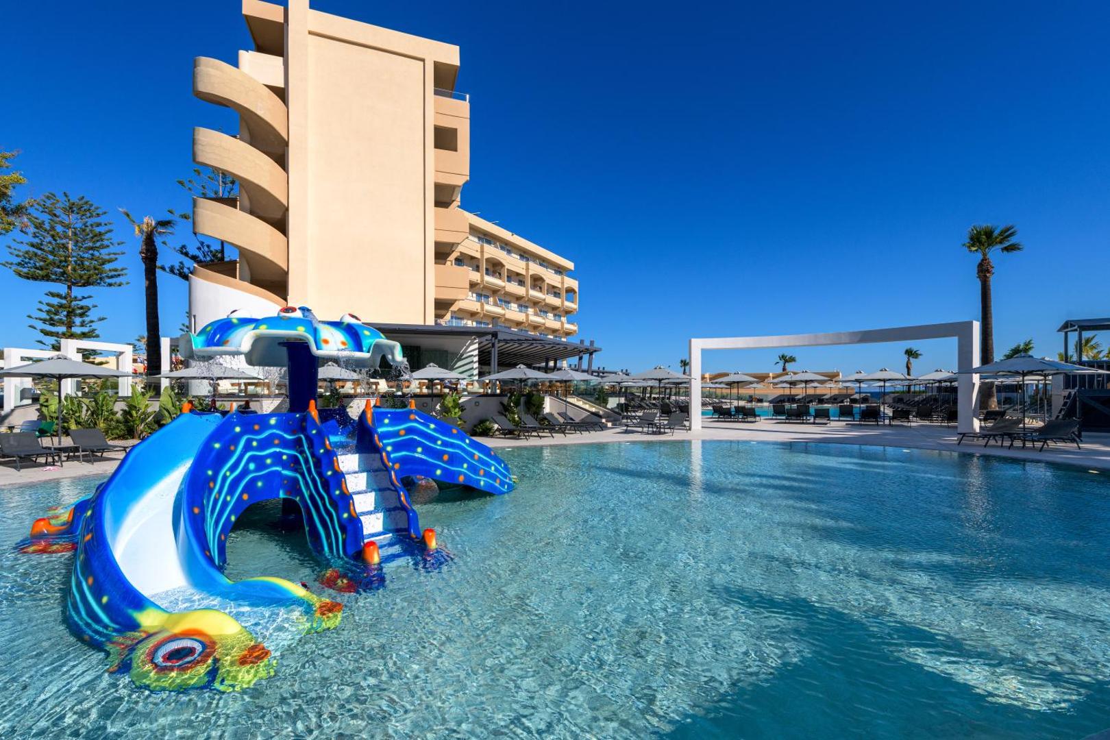 Sun Beach Resort bērnu baseins ar slidkalniņu astoņkāja formā Rodā — saulaina atpūtas zona ar sauļošanās krēsliem un viesnīcas ēku fonā.