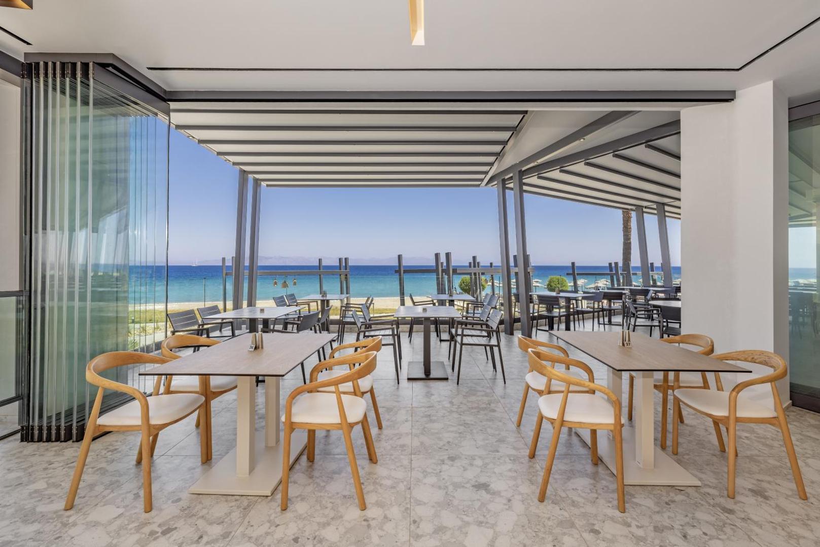 Sun Beach Resort gaišs restorāns ar panorāmas logiem un skatu uz zilo jūru Rodā — moderni koka krēsli un galdiņi.