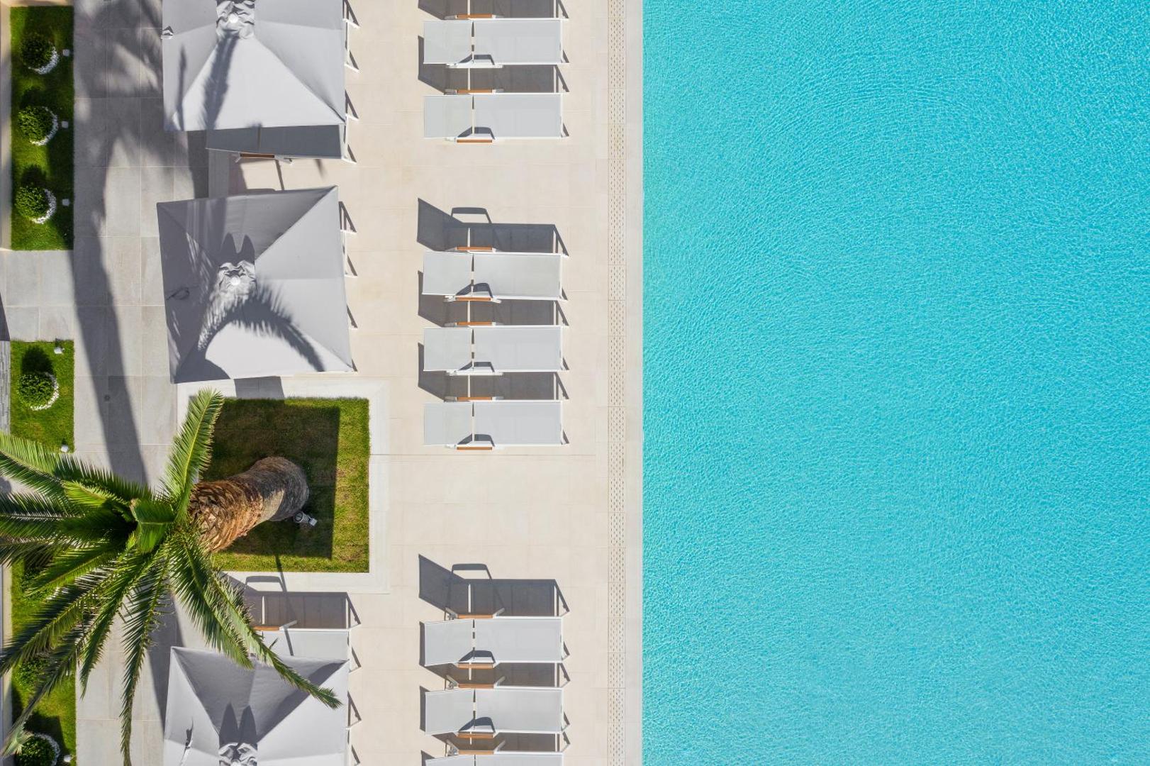 Skats no putna lidojuma uz Sun Beach Resort sauļošanās zonu ar baltiem saulessargiem, krēsliem un palmu pie baseina malas