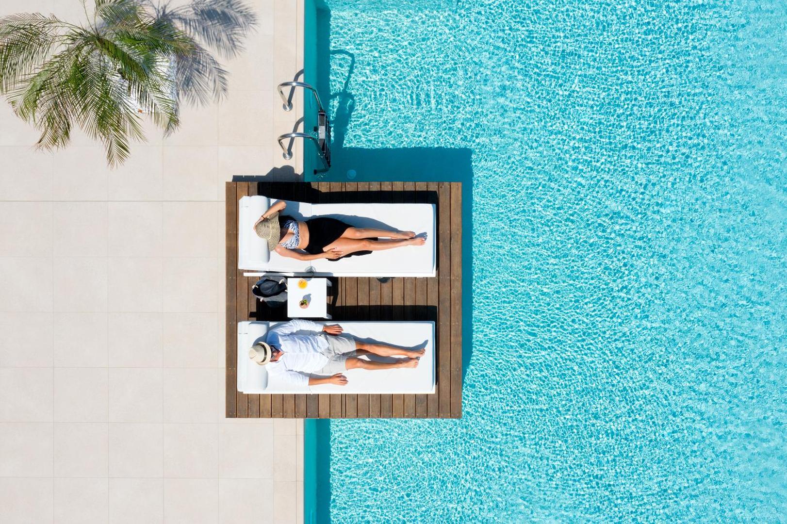 Aerofoto uzņēmums ar diviem viesiem, kuri atpūšas uz sauļošanās krēsliem Sun Beach Resort baseina vidū uz koka platformas