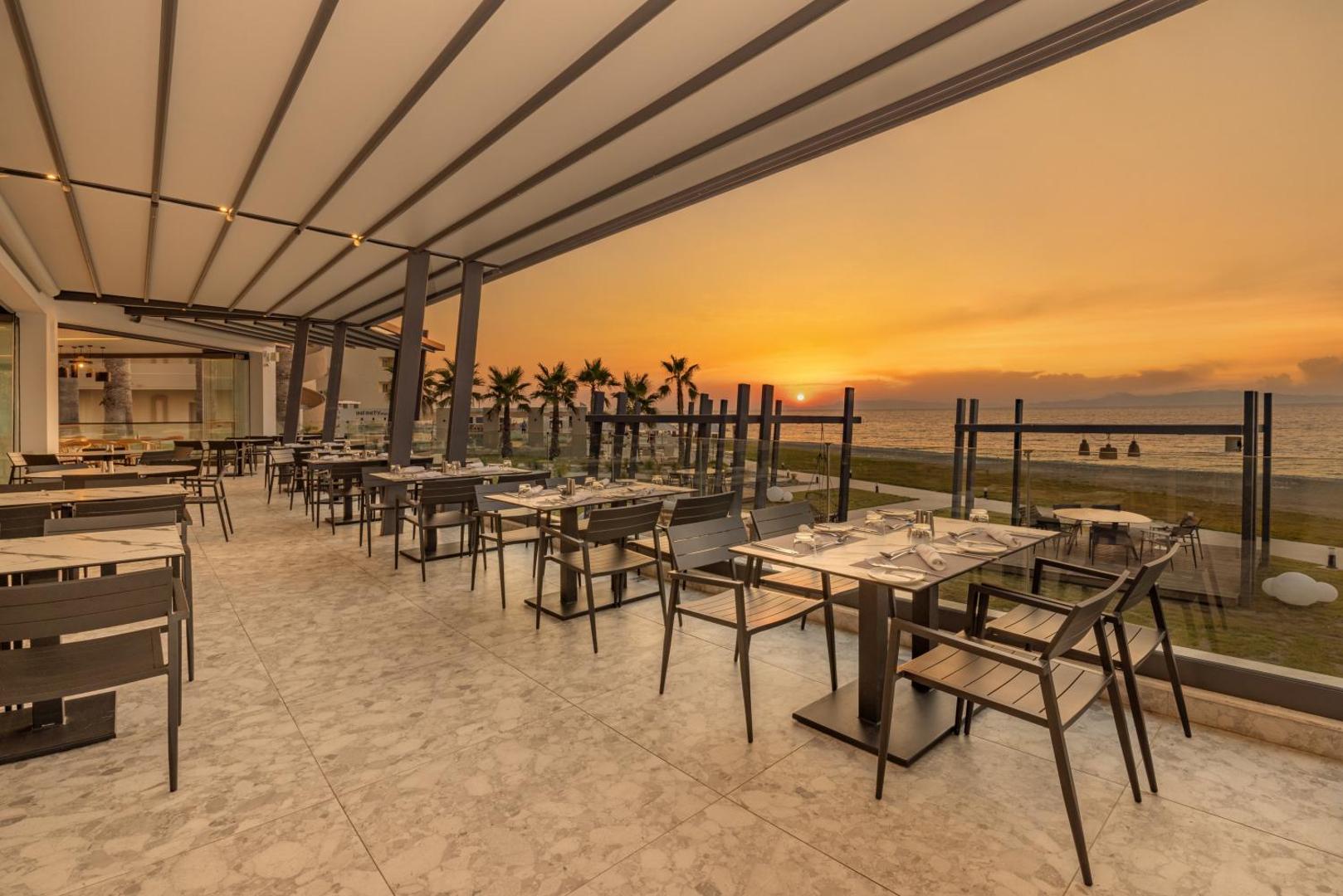 Sun Beach Resort āra terases restorāns saulrieta laikā ar skatu uz jūru Rodas salā, Grieķijā — galdiņi piekrastē zem nojumes.