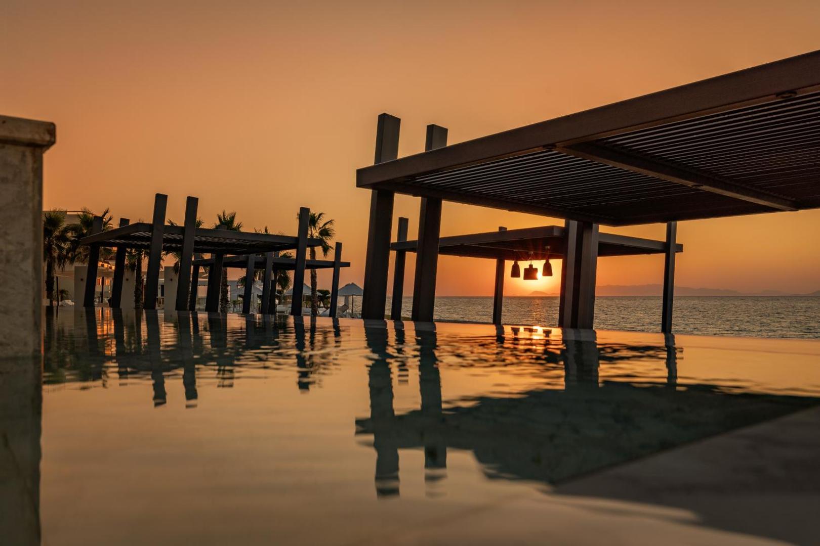 Saulriets Sun Beach Resort pie baseina ar pārplūdes malu — koka lapenes siluets pret oranžām debesīm un jūras horizontu
