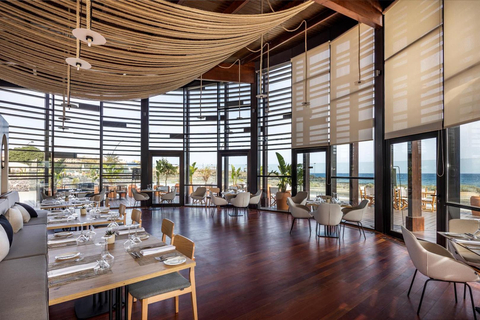 Barceló Tenerife Royal Level plašā restorāna zāle ar panorāmas logiem uz okeāna piekrasti un dabisku apgaismojumu Tenerifē