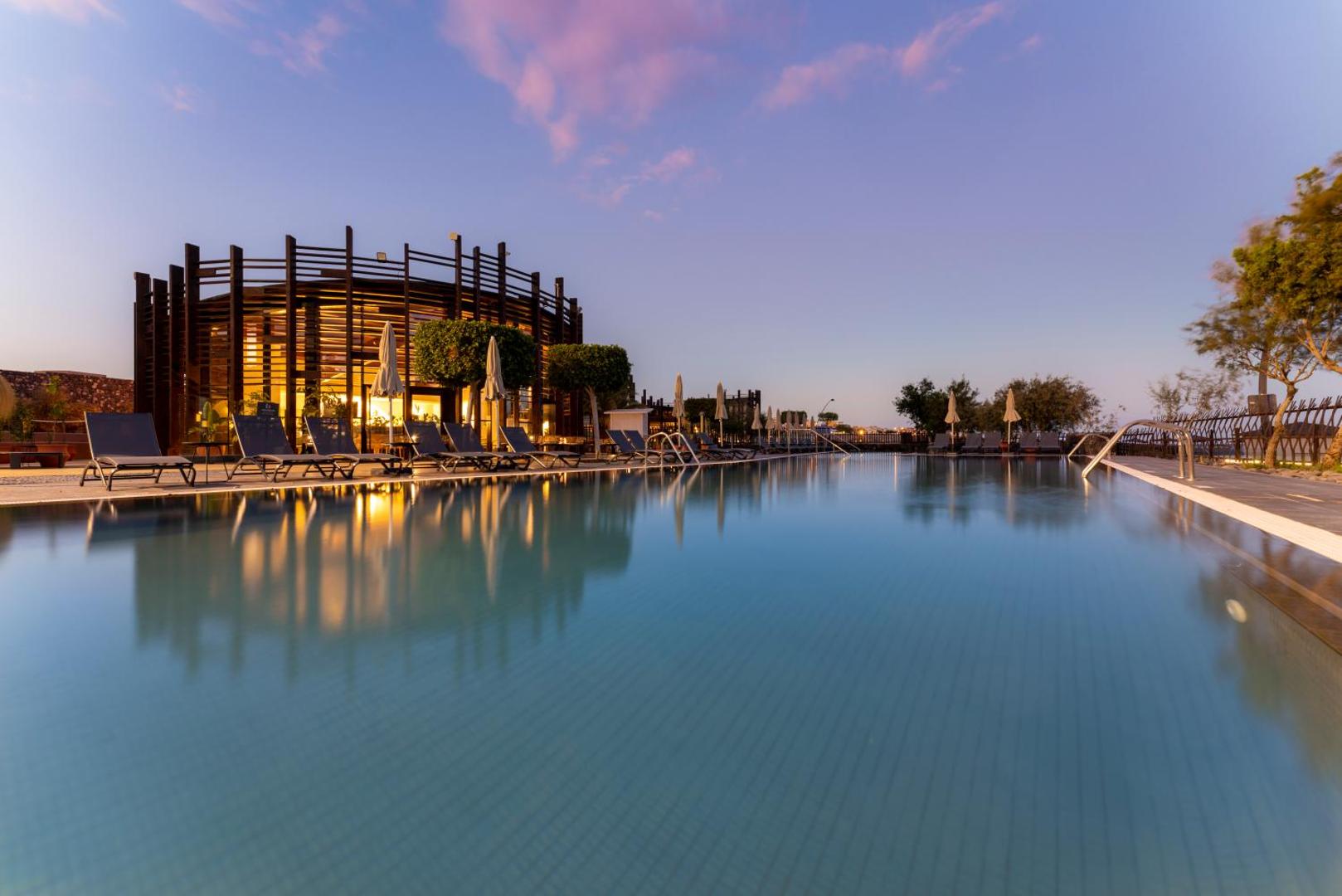 Barceló Tenerife Royal Level baseina zona saulrieta laikā ar izgaismotu ēku un atpūtas krēsliem Tenerifes piekrastē