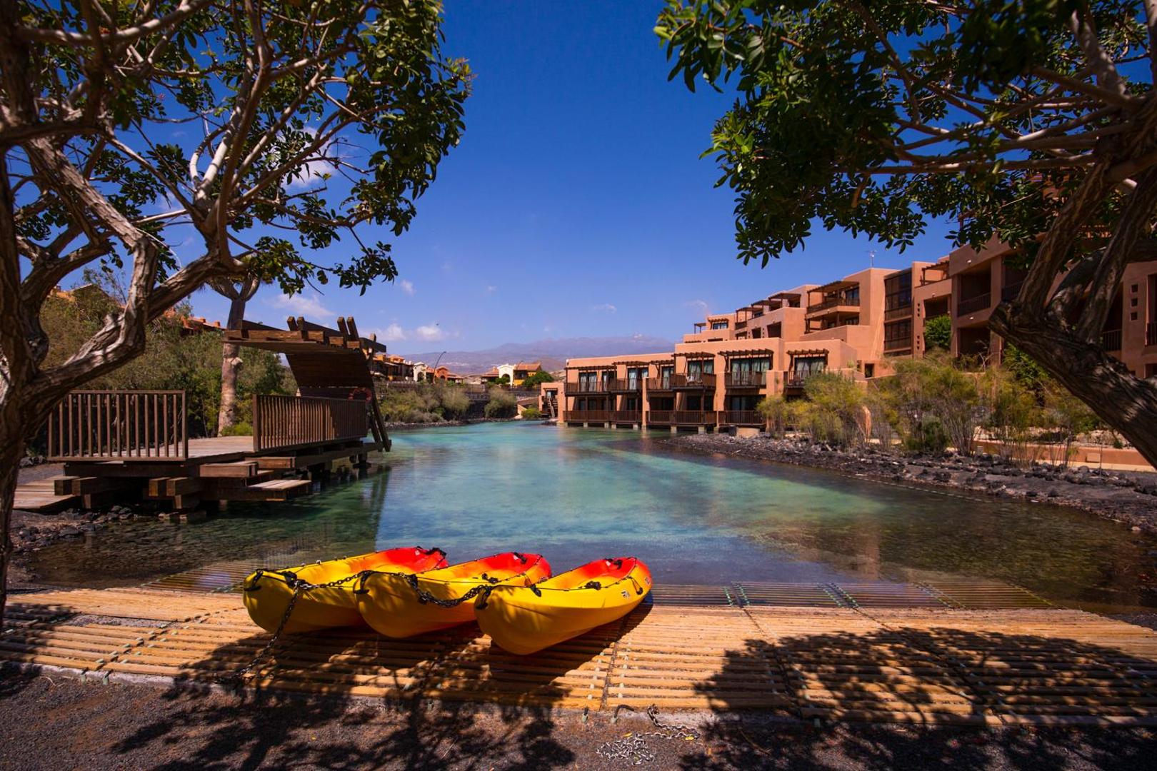 Barceló Tenerife Royal Level viesnīcas teritorija ar mākslīgo ezeru, dzelteniem kajakiem un terakotas krāsas ēkām Tenerifes piekrastē