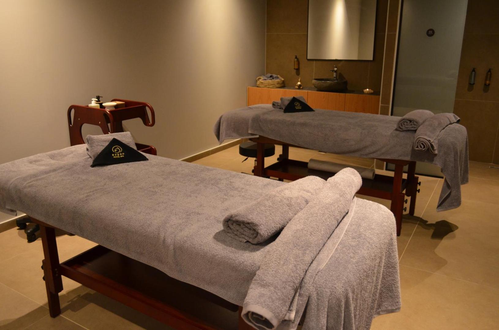 Cook's Club Ialysos Rhodes spa procedūru telpa ar diviem masāžas galdiem, pelēkiem dvieļiem un mierīgu apgaismojumu