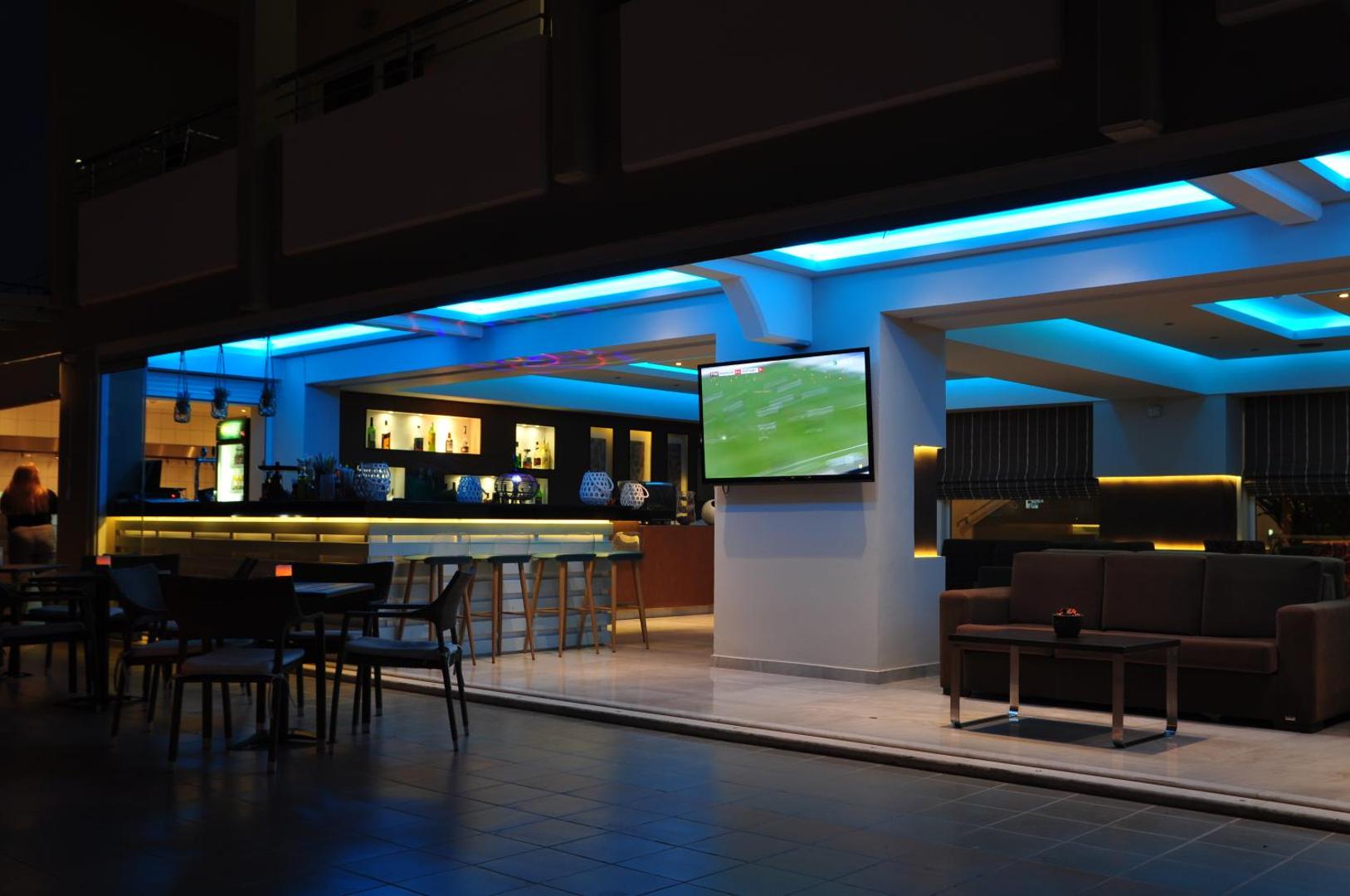 Sea Melody Beach Hotel Apartments stilīgais bārs ar zilu LED apgaismojumu, TV ekrānu un ērtiem dīvāniem atpūtai vakarā.