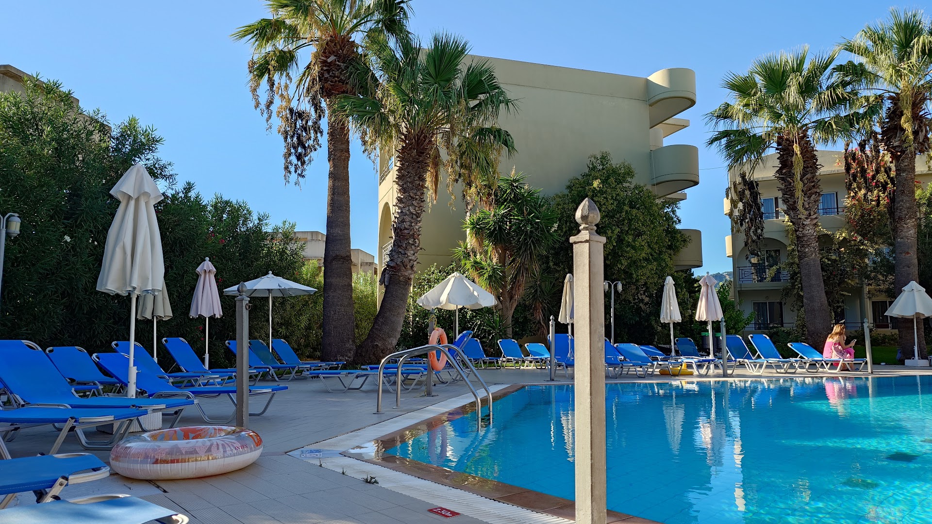 Sea Melody Beach Hotel Apartments sauļošanās krēsli un saulessargi pie baseina Rodas salā — palmu ieskauta atpūtas zona