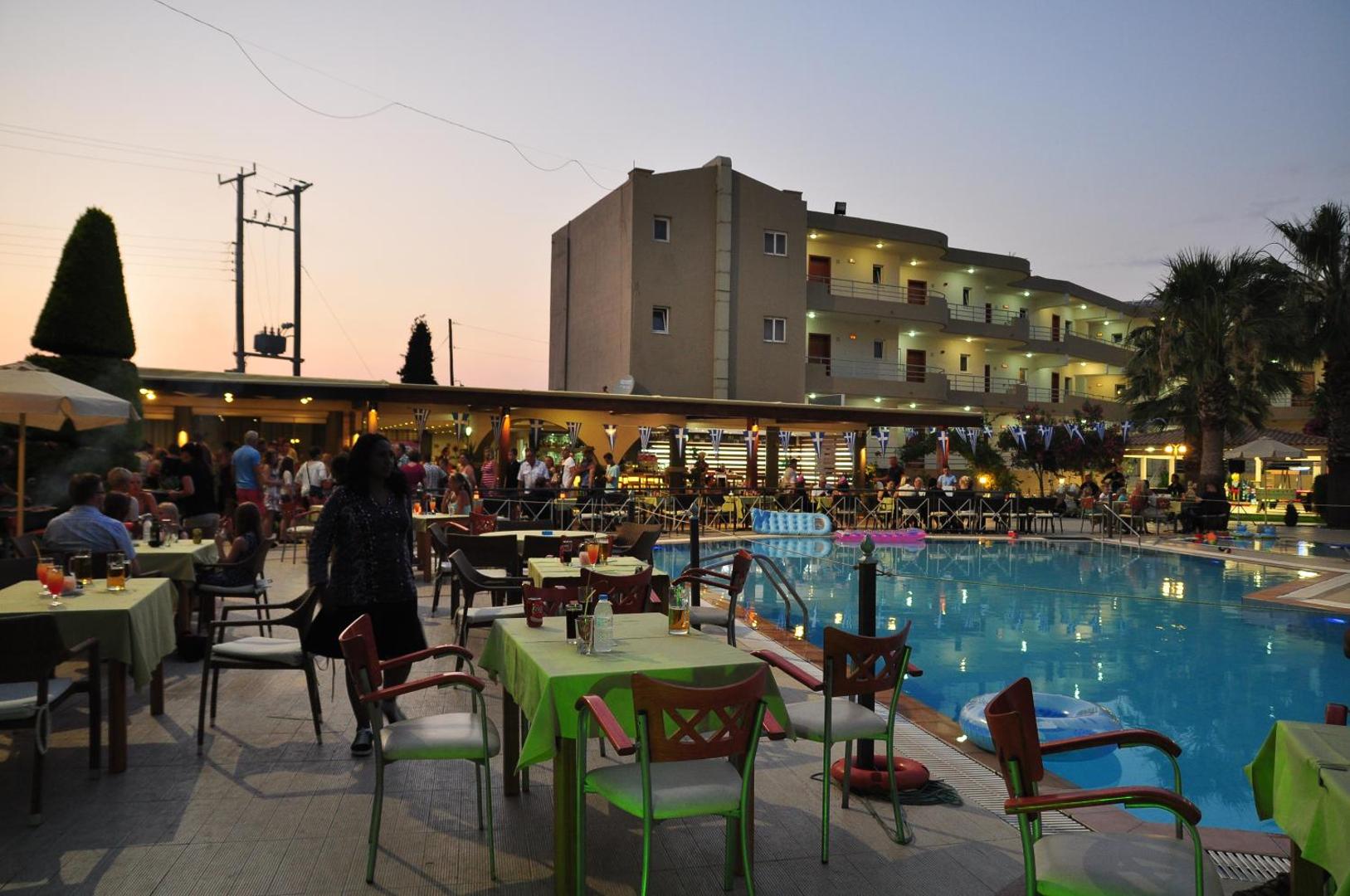 Sea Melody Beach Hotel Apartments āra baseins vakarā ar restorāna zonu, galdiņiem un viesnīcas ēku Rodas salā, Grieķijā.