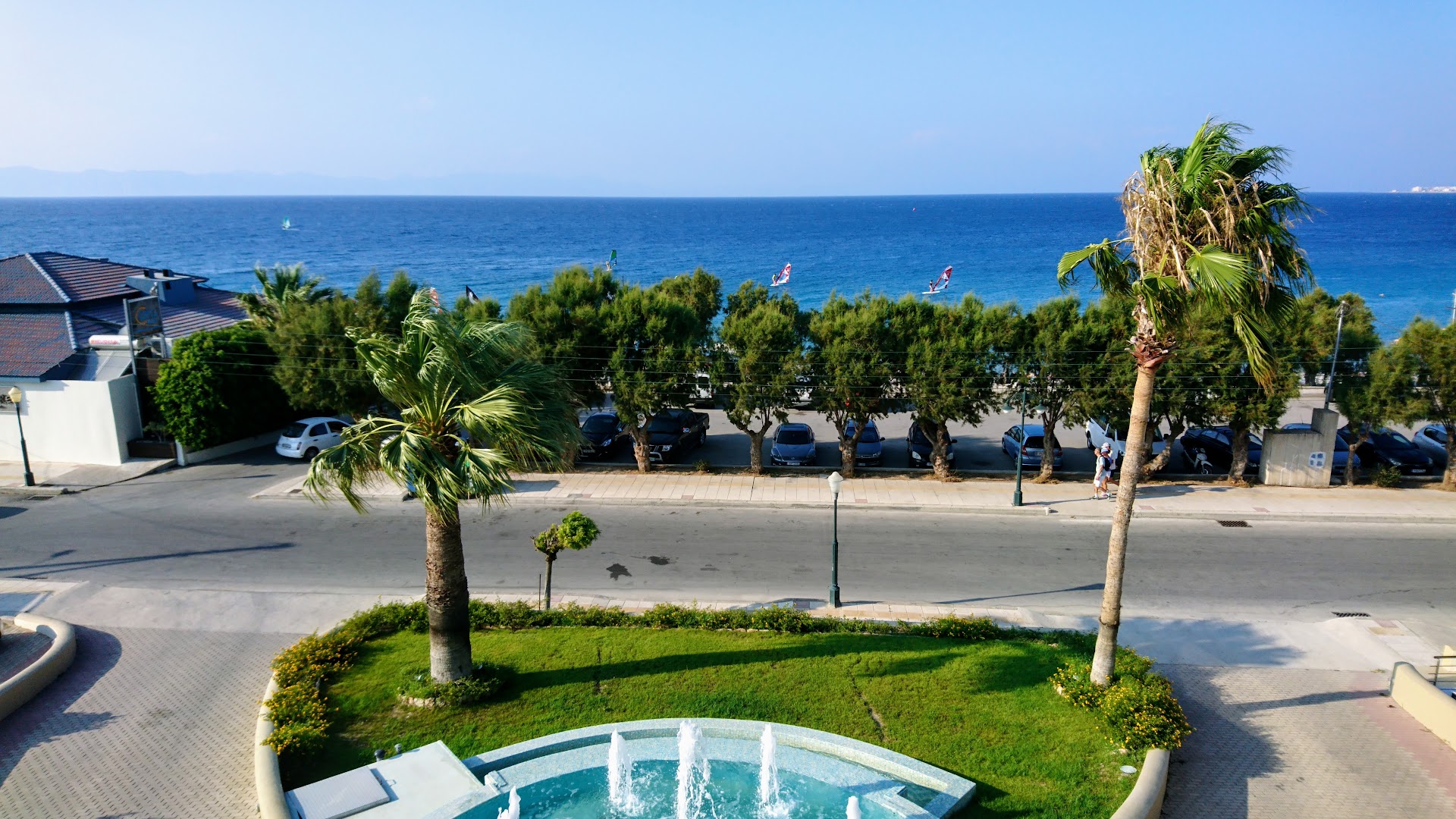 Sea Melody Beach Hotel Apartments teritorija ar strūklaku, palmām un skatu uz jūru, kur redzami vindsērfotāji Rodas piekrastē
