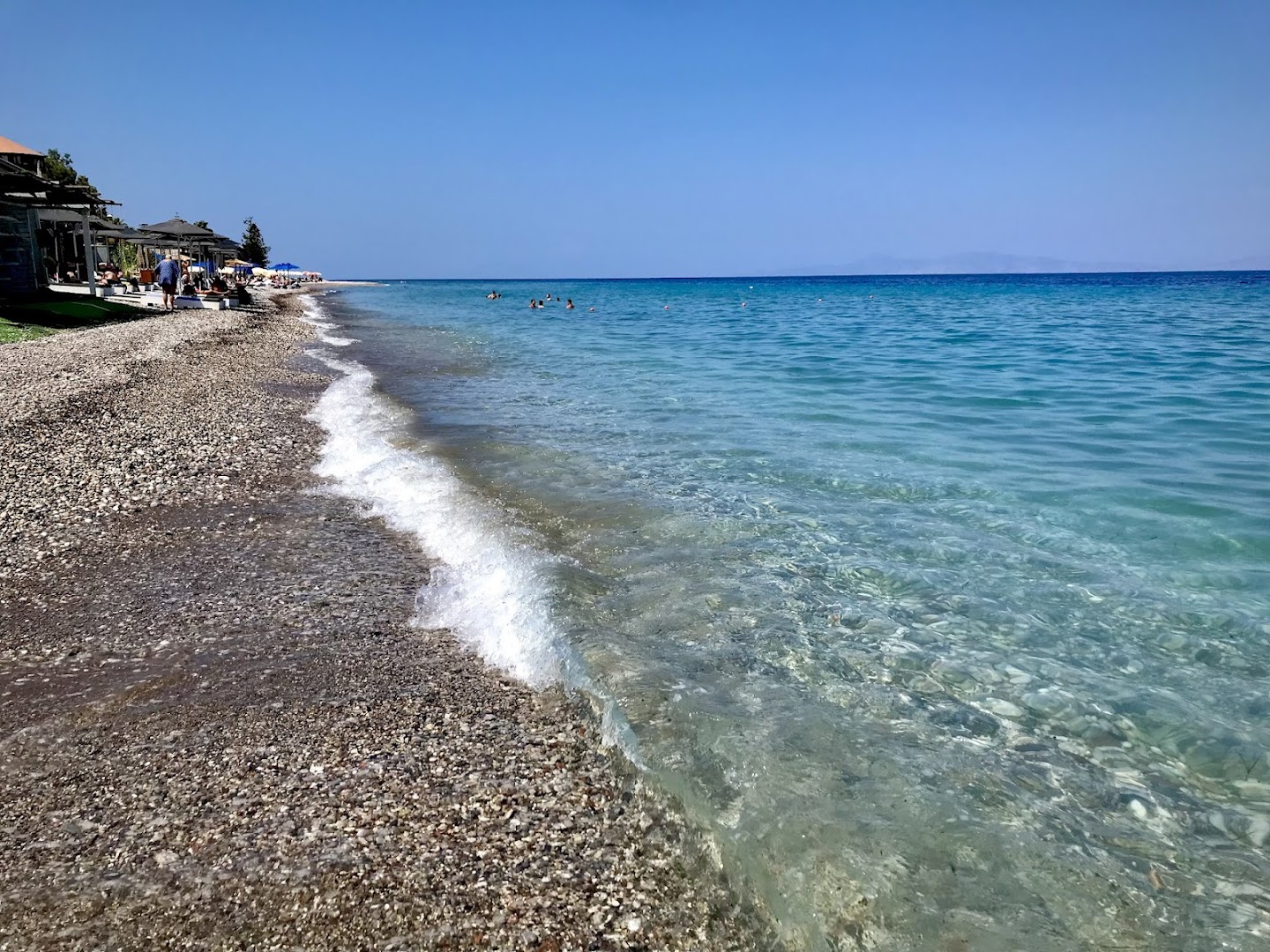 Sea Melody Beach Hotel Apartments oļu pludmale ar dzidru zilu jūras ūdeni un saulessargiem Rodas piekrastē.