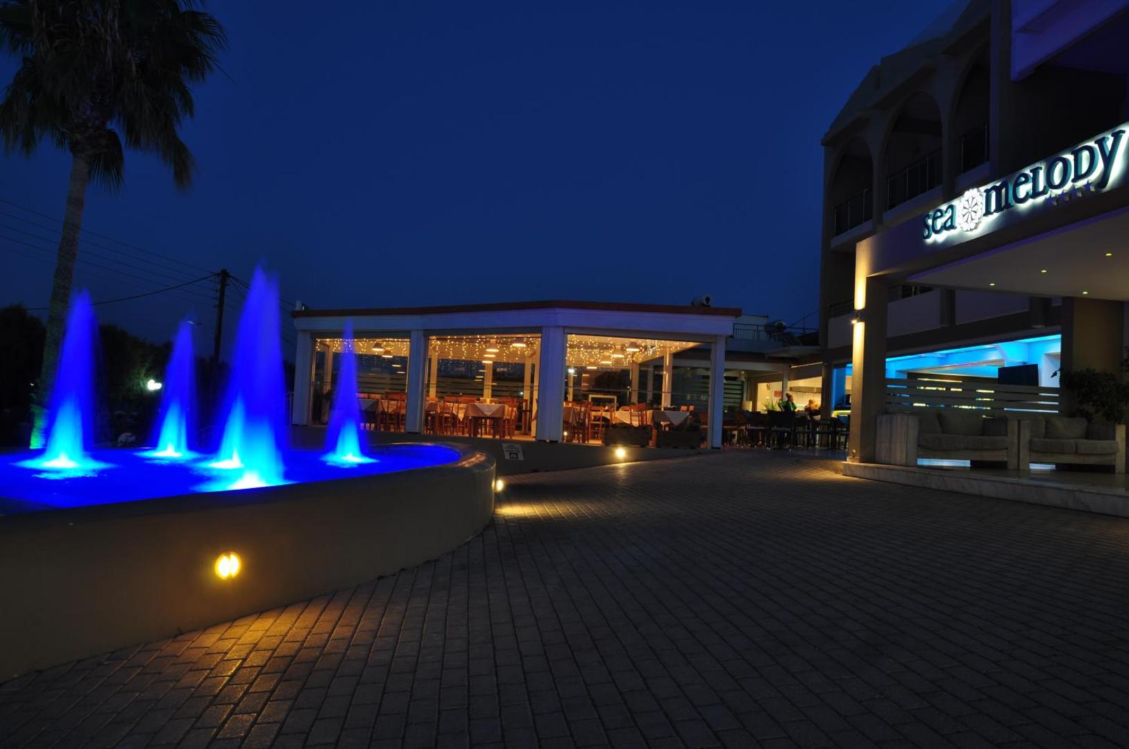 Sea Melody Beach Hotel Apartments restorāna terase un izgaismota strūklaka naktī — romantiska atmosfēra vakariņām pie jūras