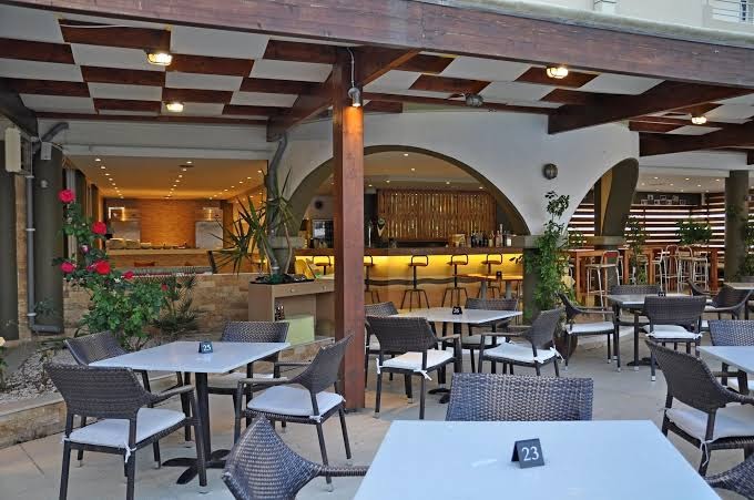 Sea Melody Beach Hotel Apartments āra terase ar modernām mēbelēm, bāru fonā un dekoratīviem augiem vakara apgaismojumā