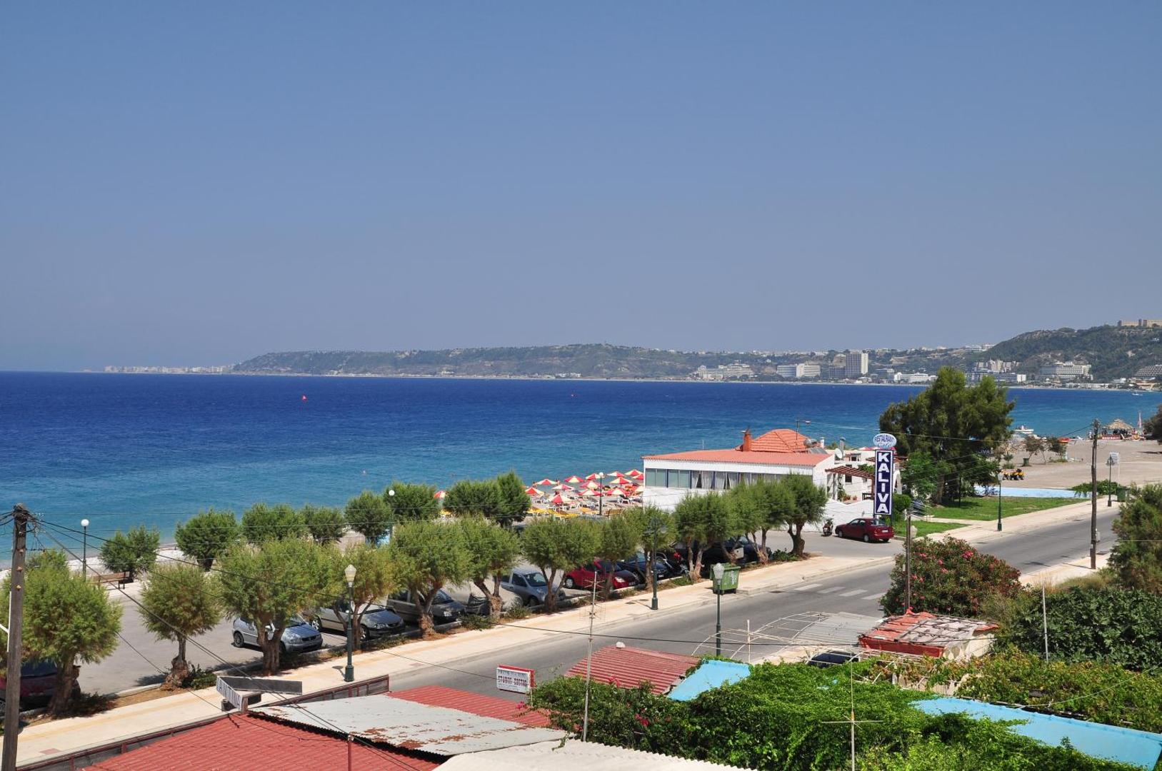 Skats no Sea Melody Beach Hotel Apartments uz Egejas jūru Rodas salā — piekrastes ceļš, koki un pludmale ar saulessargiem