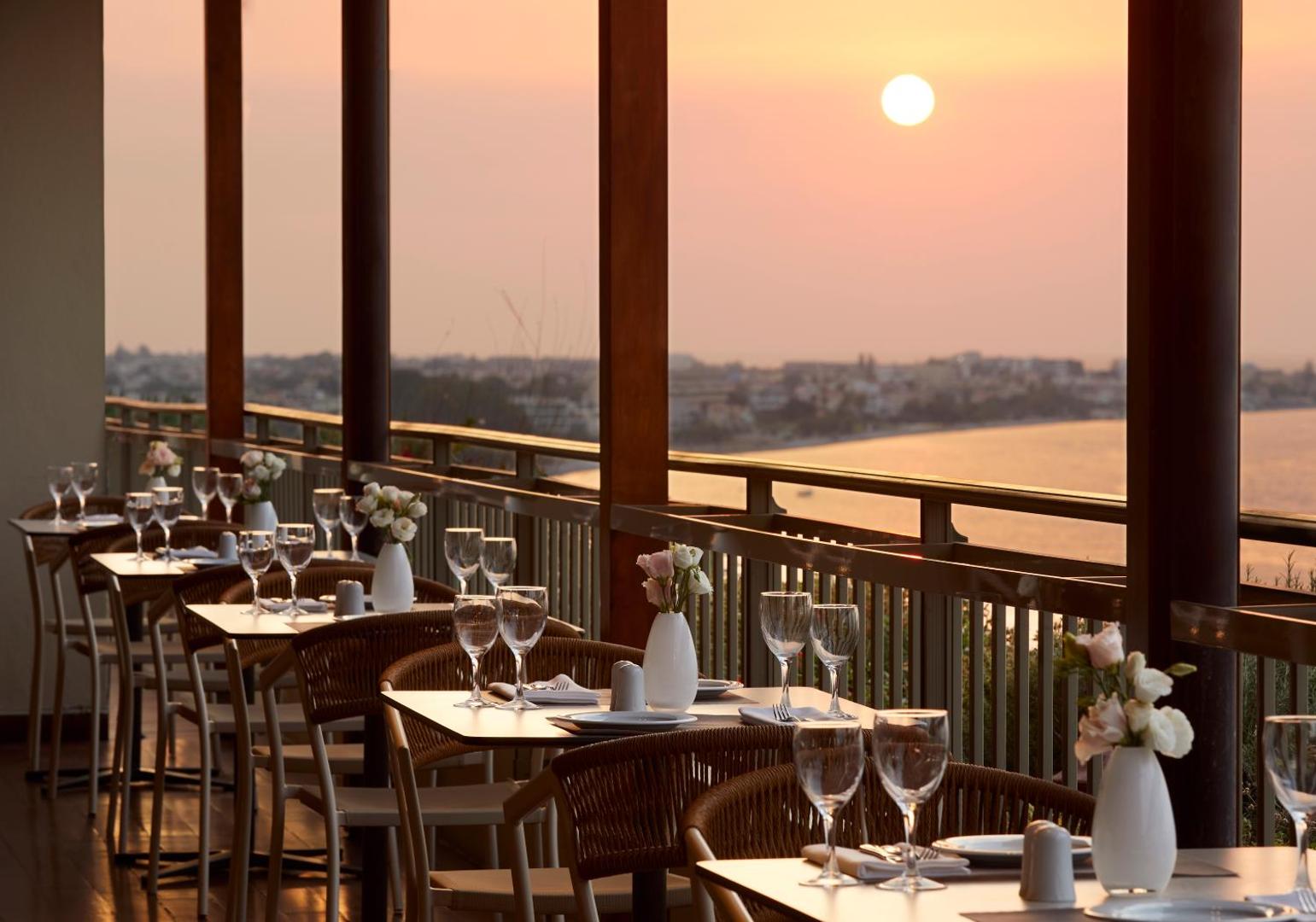 Amus Hotel & Spa restorāna galdiņu rinda pie margām ar skatu uz piekrasti un saulrietu Rodas salā