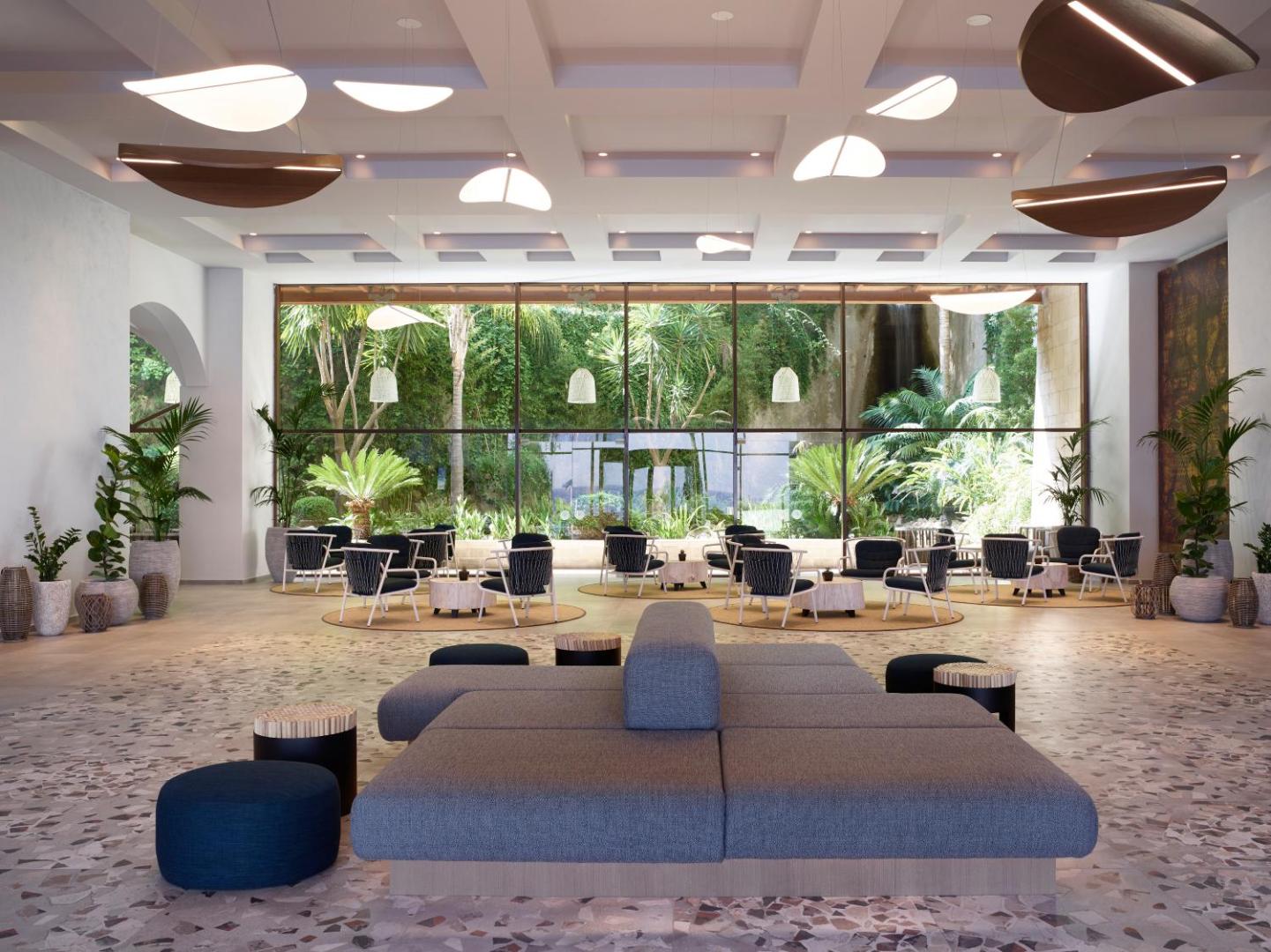 Amus Hotel & Spa plašais vestibils ar modernām mēbelēm, dizaina gaismekļiem un lielu panorāmas logu uz dārzu Budvā, Melnkalnē
