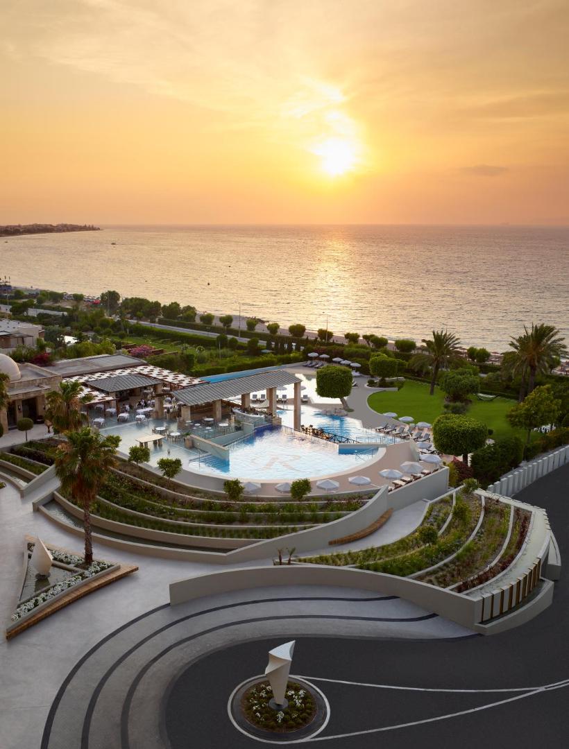 Amus Hotel & Spa skats no augšas uz baseinu kompleksu un dārzu saulrieta laikā pie Egejas jūras krasta.