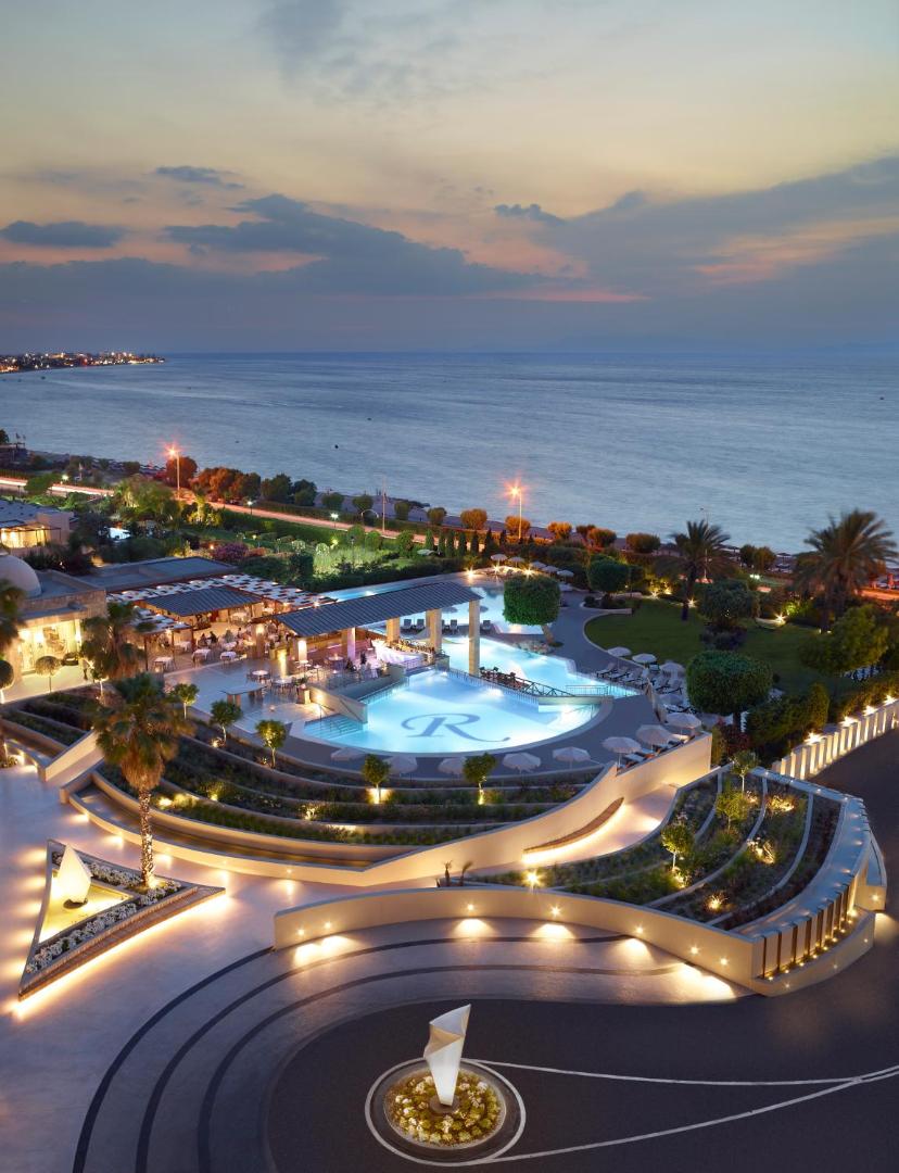 Amus Hotel & Spa izgaismota teritorija vakarā ar skatu uz baseinu, restorāna zonu un jūras horizontu krēslas stundā.