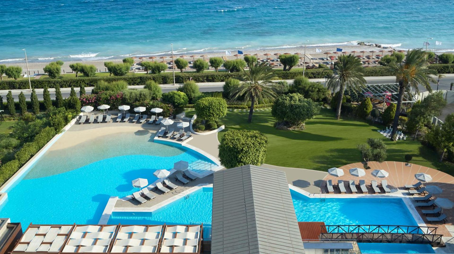 Amus Hotel & Spa āra baseinu zona ar sauļošanās krēsliem, zaļu zālienu un skatu uz jūras piekrasti saulainā dienā