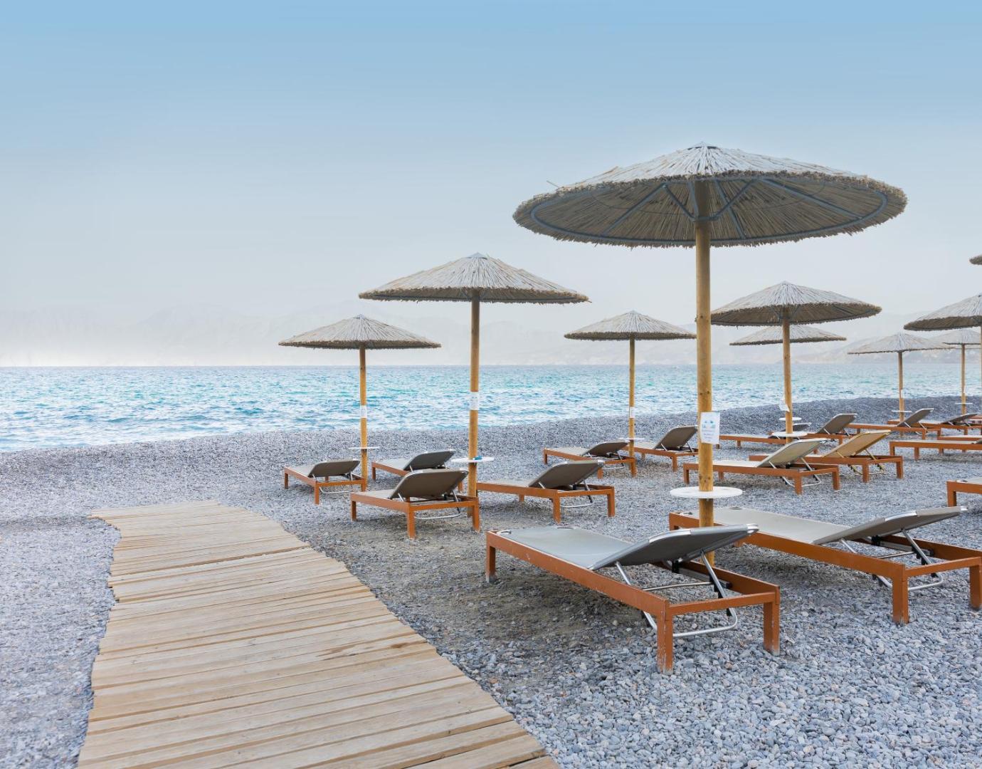 Ariadne Beach Hotel Malia pludmales zona ar koka laipām, moderniem zviļņiem un salmu saulessargiem uz oļu seguma pie jūras