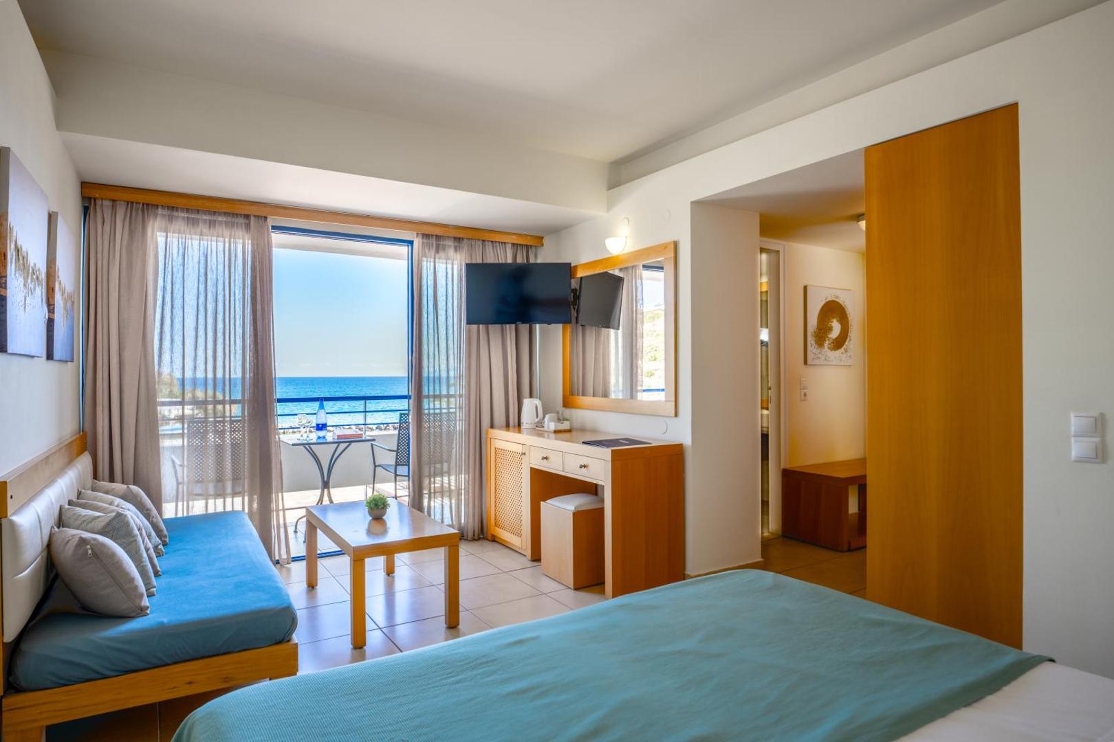 Skats no Ariadne Beach Hotel numura uz jūru un balkonu Malijā — numurā ir rakstāmgalds, televizors un zils dīvāns pie loga.