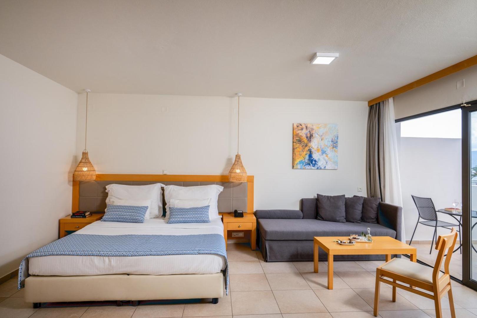 Ariadne Beach Hotel numurs ar divguļamo gultu, pelēku dīvānu un balkonu Malijā — interjers ar koka elementiem un pītiem gaismekļiem.