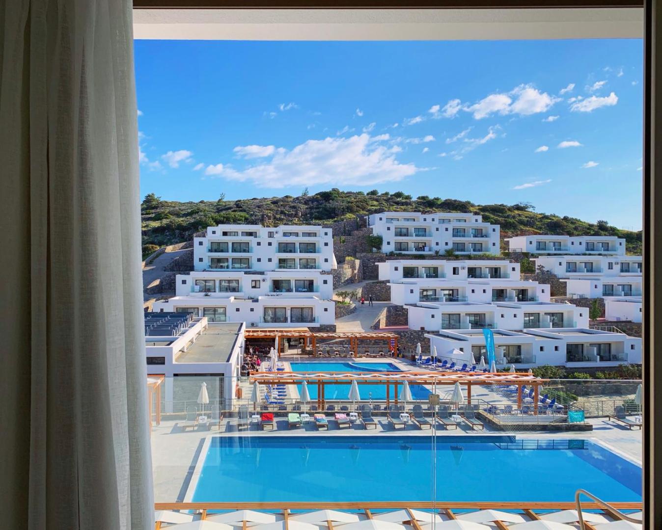 Skats pa logu uz Ariadne Beach Hotel teritoriju Malijā — redzami vairāki baseini, sauļošanās terases un viesnīcas korpusi kalna nogāzē.