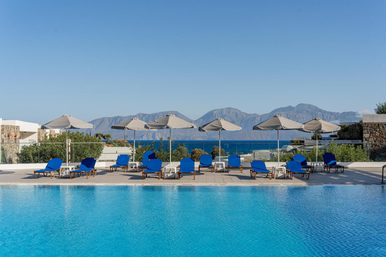 Ariadne Beach Hotel baseina mala ar zilajiem sauļošanās krēsliem un saulessargiem uz kalnu un jūras fona