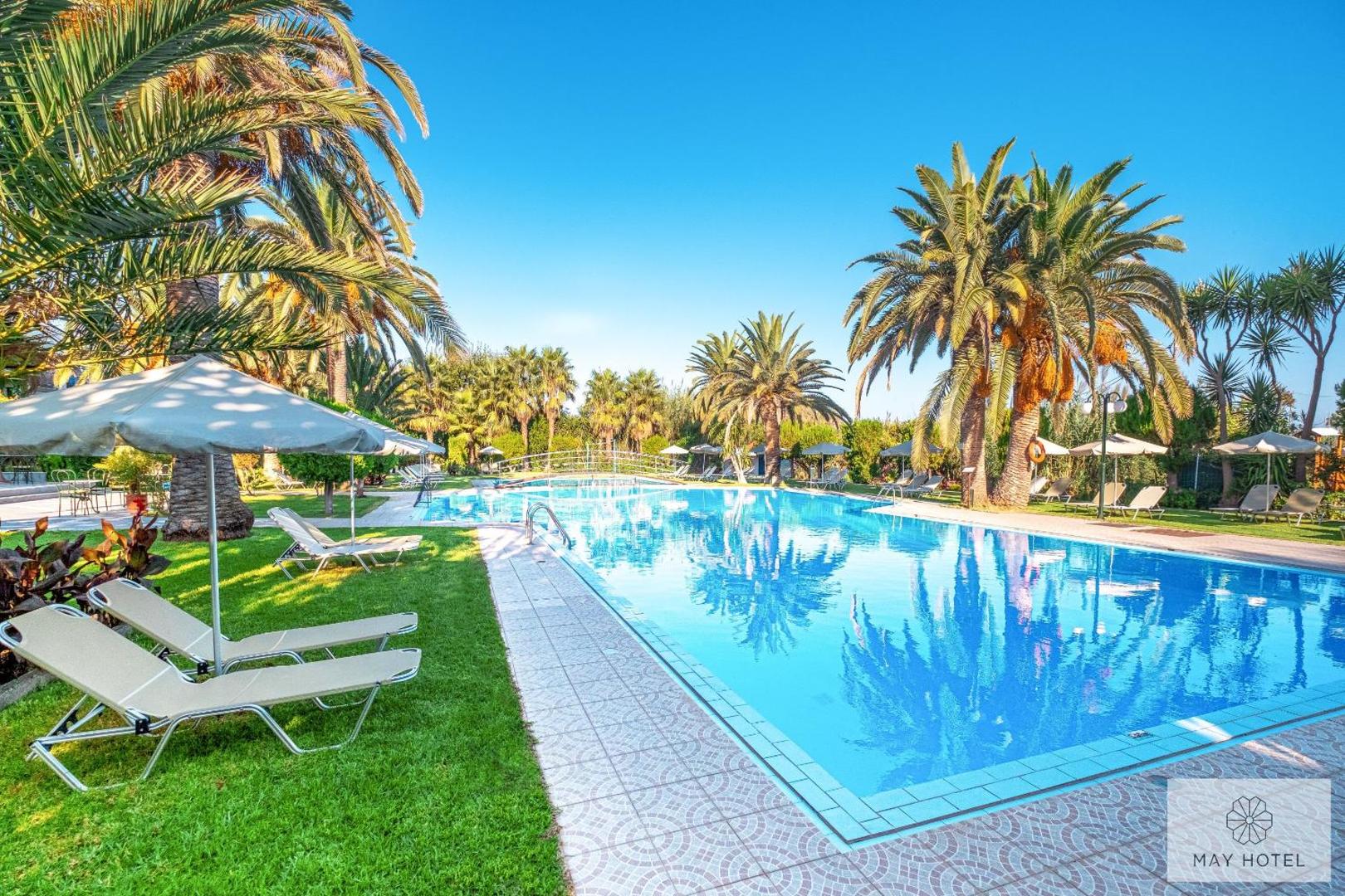 May Hotel atpūtas zona pie baseina ar ērtiem zviļņiem uz zaļa zālāja un lielām palmām Krētas kūrortā.