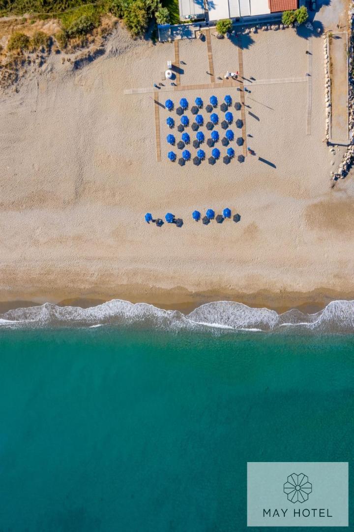 Aerofoto skats uz May Beach Hotel privāto pludmali Krētā — zili saulessargi uz smilšu fona un dzidrs jūras ūdens