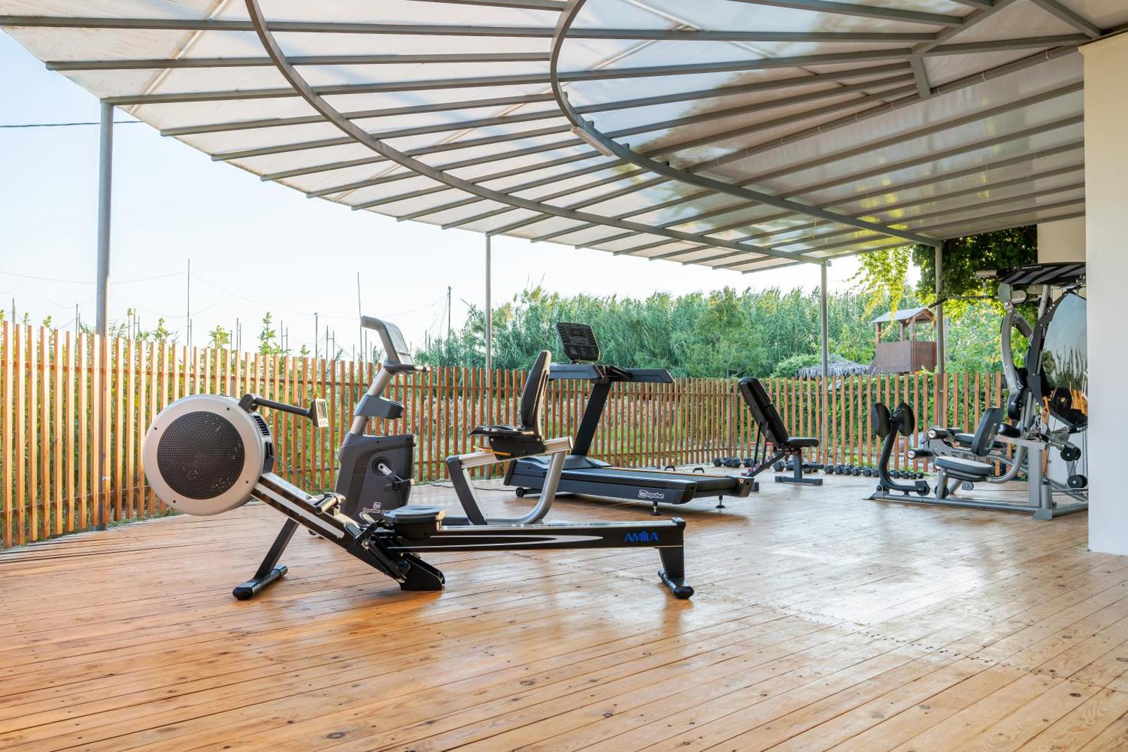 May Hotel āra fitness zona zem nojumes Krētā — trenažieri, airēšanas trenažieris un skrejceliņš uz koka terases