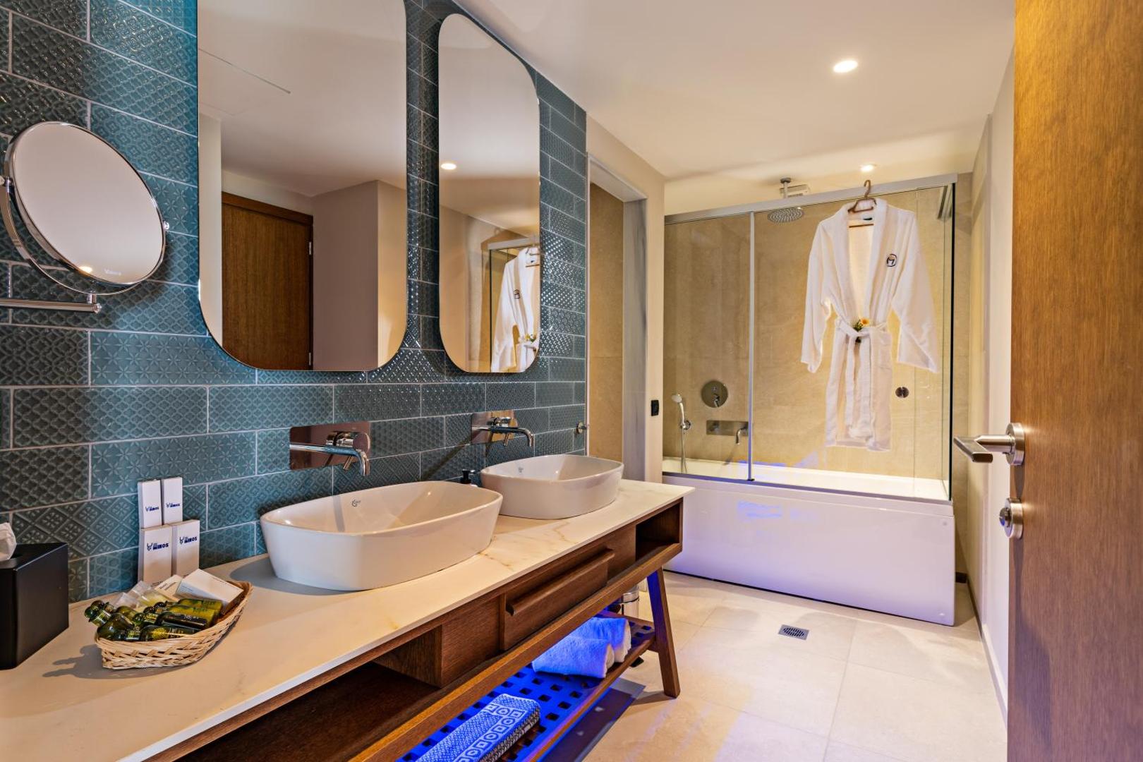 Minos Ambassador Suites & Spa moderns vannas numurs ar divām izlietnēm, lielu spoguli un vannu aiz stikla sienas