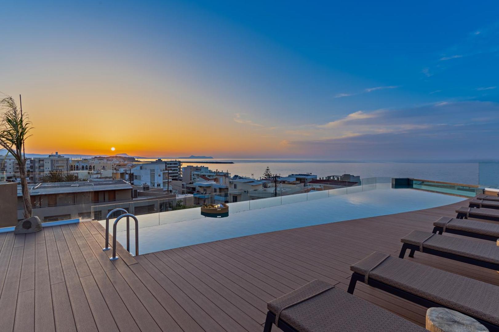 Minos Ambassador Suites & Spa jumta terase ar baseinu ar pārplūdes malu, sauļošanās krēsliem un saulrietu pār jūru